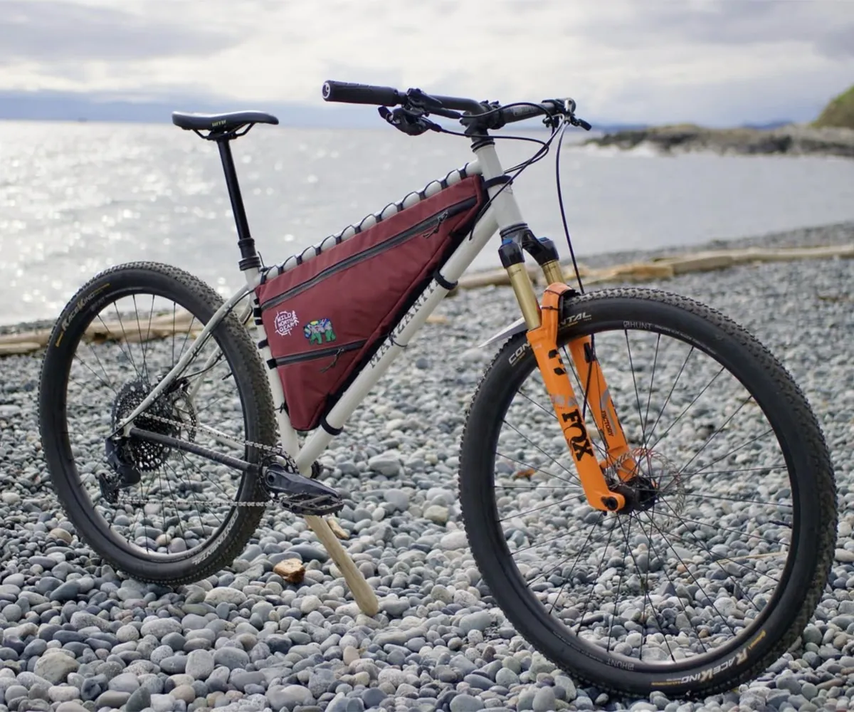A hardtail bike on a pebble beach with a burgundy Wild North Gear frame bag